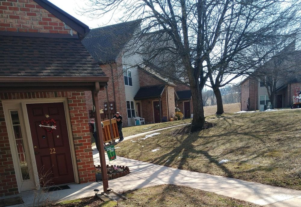 Two neighbors bringing an upright piano down the walkway on a dolly, heading for an apartment door.