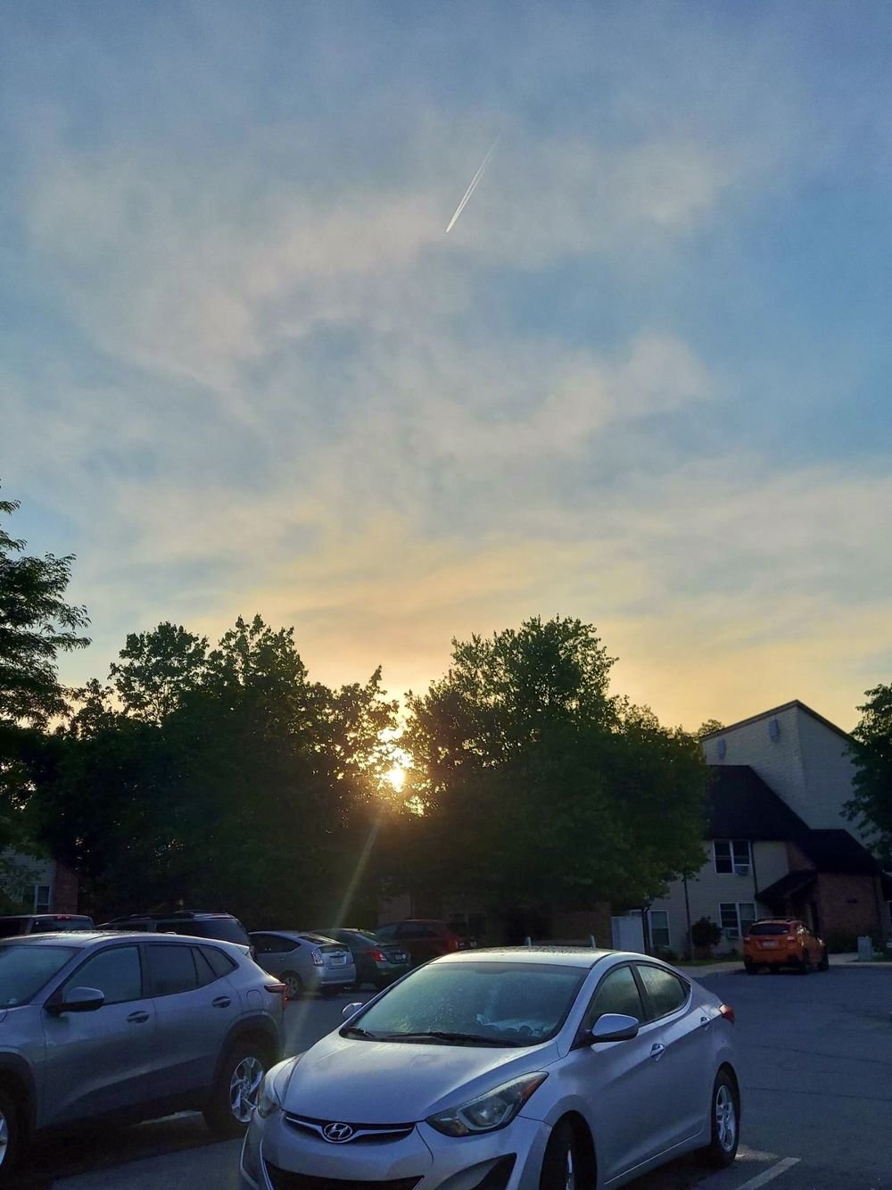 The sun rising through trees above the parking lot, with an apartment building at the right.